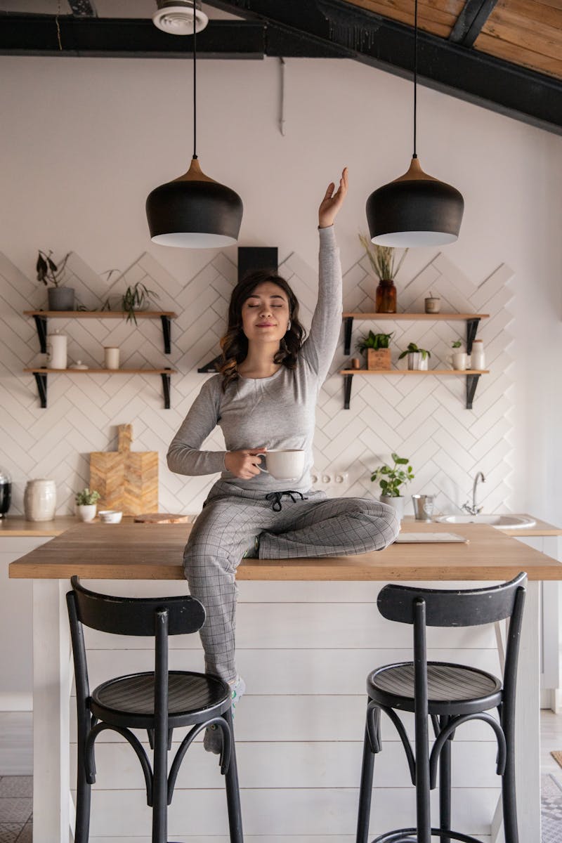 Dreamy young ethnic female with closed eyes in pajamas sitting with crossed legs on kitchen table with cup of hot drink and raised hand